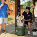 Three men at an outdoor gathering around a keg and cooler, one pouring beer into a cup while another holds a drink, under a wooden pavilion.