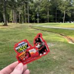 Hand holding a red promo card reading 'Who's Your Caddy?' with a cartoon caddy and food items, in a park setting (grassy field).