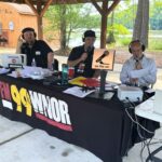Three men with headsets broadcast a live outdoor radio show from a wooden pavilion, microphones and equipment on a long table covered with a banner, with a sunny lake backdrop and an 'on the air' sign in the foreground.