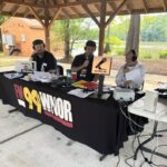 Three men wearing headphones sit at a broadcast table under a wooden pavilion, speaking into microphones with an 'on the air' sign nearby.