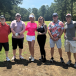 Six people standing on a golf course fairway posing for a group photo on a sunny day with trees in the background and a golf cart partly visible to the right.