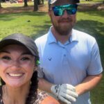 Two friends pose for a sunny outdoor photo on a golf course; woman in the foreground with a braid and tattoo, man behind wearing a cap, reflective sunglasses, polo shirt and golf glove.