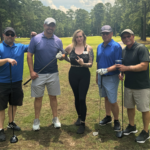 Five people posing together on a sunny golf course, each with a club, smiling at the camera.