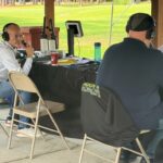 Two men wearing headphones sit at a broadcast table under a pavilion; one holds a microphone while the other reads notes, with laptops and gear nearby (outdoors).