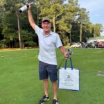 Smiling man in a light polo and navy shorts on a golf course, raising a champagne bottle while carrying a Virginia Natural Gas tote bag.
