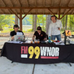 Three men with headphones broadcasting at an outdoor park pavilion, microphones and audio gear on a table covered with an FM99 WNOR banner.