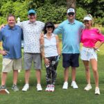 Five friends standing on a sunny golf course with arms around each other, smiling at the camera.
