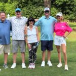 Five adults posing together on a sunny golf course, arms around each other and smiling.