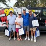 Four adults posing in front of a black FM99 van, smiling and holding white gift bags.
