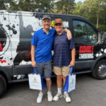 Two men stand side by side in front of a branded van, smiling, each holding a blue and white tote bag in a casual outdoor setting.