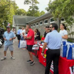 Group of people gathered around a table with blue and white gift bags in an outdoor event near a building, with trees in the background.