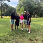 Five golfers posing together on a sunny golf course, smiling and in casual golf attire.