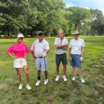 Four golfers pose on a sunny golf course, with a woman in a bright pink polo shirt and three men in casual beige/white shirts.