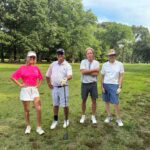 Four golfers pose on a sunny, green golf course with trees in the background, smiling at the camera.