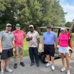 Five adults posed by two beige golf carts on a sunny golf course, smiling at the camera with green trees in the background.