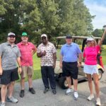 Group of five adults standing beside golf carts on a sunny course, posing for a photo.