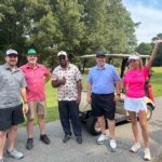 Five adults pose for a photo by golf carts on a sunny golf course; the woman in pink raises her arm in celebration.
