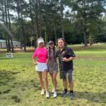Three friends pose together on a grassy area with tall trees in the background, smiling at the camera.
