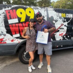 Two people posing in front of a FM99 radio van with splatter graphics; one person holds a bag.