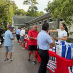 People line up at an outdoor community event, volunteers at a table handing out blue and white bags to attendees on a sunny day.
