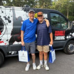 Two men stand side by side in front of a graphic van, smiling, each holding a blue event bag in a parking lot.