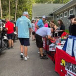 People browse booths at an outdoor streetside event; tents and banners line a walkway, with attendees looking at items and chatting.