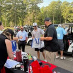 Group of people gathered around a table with items at an outdoor event, volunteers helping attendees in a park setting.