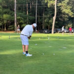 Man in light clothing stands on a grassy golf practice area, mid-swing with a club, with trees and other players visible in the background.