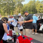 People gather around a registration table with red swag bags and bottles at an outdoor event, with golf carts and trees in the background.