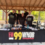 Six staff members stand behind a table with an FM 99 WNOR banner at an outdoor event under a wooden pavilion.