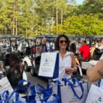 Woman in sunglasses at an outdoor event, standing behind a table with blue gift bags and a white tote bag in hand.