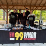 Five men stand behind a black FM99 WNOR booth table at an outdoor pavilion, with gear and swag displayed.