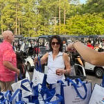 Woman in white top and sunglasses picks a blue-and-white tote bag at an outdoor event; a man in a pink shirt stands nearby.