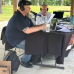 Two men recording a podcast outdoors under a pavilion; one speaks into a microphone, the other smiles at papers on the table.