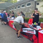 People line up at outdoor vendor tables with red-and-black banners reading 'HAMPTON ROADS CLASSIC ROCK' outside a stone building tall trees in the background.