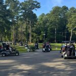 Cluster of golf carts parked in a sunny lot beside a pine forest, players loading gear.