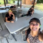 Smiling woman in leopard print top takes a selfie at outdoor pavilion with three others seated at long tables and drinks on the table.