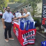 People at an outdoor event exchange a handshake across a table covered with a red FM99 Rocks Hampton Roads banner and gift bags on display.