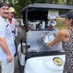 People gather around a golf cart booth in a park; a woman in a leopard-print dress hands a flyer to a man in a white cap inside the cart window.