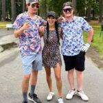 Three friends pose together in a park: two men in floral shirts flanking a woman in a leopard-print dress, all smiling and wearing casual summer outfits.