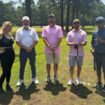 Five adults posing on a golf course with clubs, ready to play and smiling at the camera.