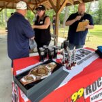 Award table under a park pavilion with a championship belt in a box and multiple trophies on display as people chat nearby