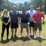 Five people standing together on a sunny golf course, posing for a group photo on the fairway with trees in the background.
