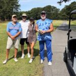Four adults posing on a sunny golf course next to a golf cart, smiling at the camera.