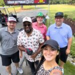 Five people pose for a selfie at an outdoor event under a red-and-black canopy; woman in a leopard-print top foreground, others in casual golf attire.