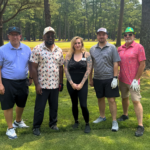 Five adults posing for a photo on a sunny golf course with trees in the background, wearing casual golf attire.