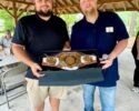 Two men pose under a wooden pavilion, presenting a boxed gold championship belt.