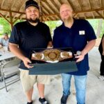 Two men pose under a wooden pavilion, presenting a boxed gold championship belt.