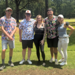 Five people posing together on a sunny golf course, wearing colorful tropical shirts and caps/visors.