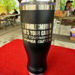 Black insulated tumbler with large silver text advertising a golf tournament sits on a red tablecloth under a wooden pavilion at a picnic area.
